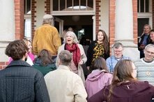 Eine Gruppe an Menschen kommt an einem Gebäude an. Die meisten sind dem Eingang zugewandt. Einige Personen schauen Richtung Kamera. Eine Person mittig lacht erfreut.