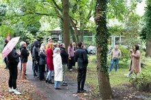 Eine Gruppe an Menschen steht draußen auf einem Weg. Sie sind umgeben von Bäumen und Rasen. Zwei Personen tragen etwas vor und stehen vor der Gruppe auf dem Rasen.