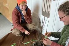 Zwei Menschen hocken über einer Decke, die auf dem Boden liegt. Auf der Decke sind Zweige eines Apfelbaumes zu sehen. Ein Bäumchen steht in einem Topf in der Mitte der Menschen. Eine Person arbeitet mit den Zweigen. Sie veredeln sie.