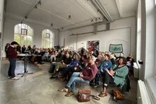 Viele Menschen sitzen auf Stühlen, die zu einer stehenden Person gerichtet sind. Sie hören der stehenden Person zu. Diese hält einen Vortrag. Im Hintergrund stehen weitere Menschen. Es wird etwas an die Wand gebeamt, was ein Beamer im Hintergrund zeigt.