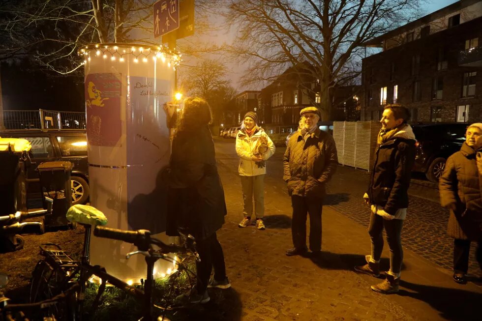 Links im Bild steht eine beleuchtete Litfaßsäule, auf der einige bunte Plakate mit Text kleben. Eine Frau beschreibt eines der bunten Plakate mit einem Stift. Rechts daneben stehen drei ältere und eine jüngere Person, die ihr dabei zuschauen. Im Hintergrund sind beleuchtete Wohnhäuser zu sehen.