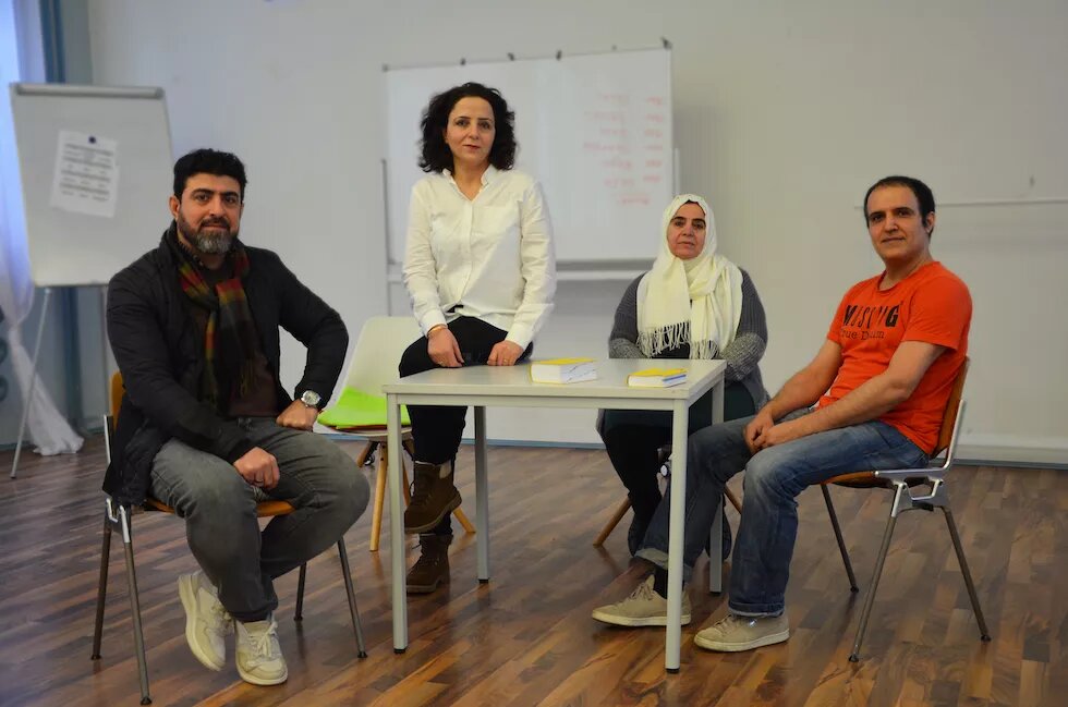 Vier Personen sind auf dem Bild zu sehen. Sie formieren sich um einen Tisch. Links sitze ein Mann mit grauem Bart und dunklen Haaren aus einem Stuhl. Rechts von ihn sitzt eine Frau mit dunklen Haaren auf dem Tisch. Rechts von ihr sitzt eine Frau mit weißem Kopftuch auf einem Stuhl. Neben ihr sitzt ein Mann mit dunklen Haaren. Im Hintergrund ist eine Tafel zu sehen