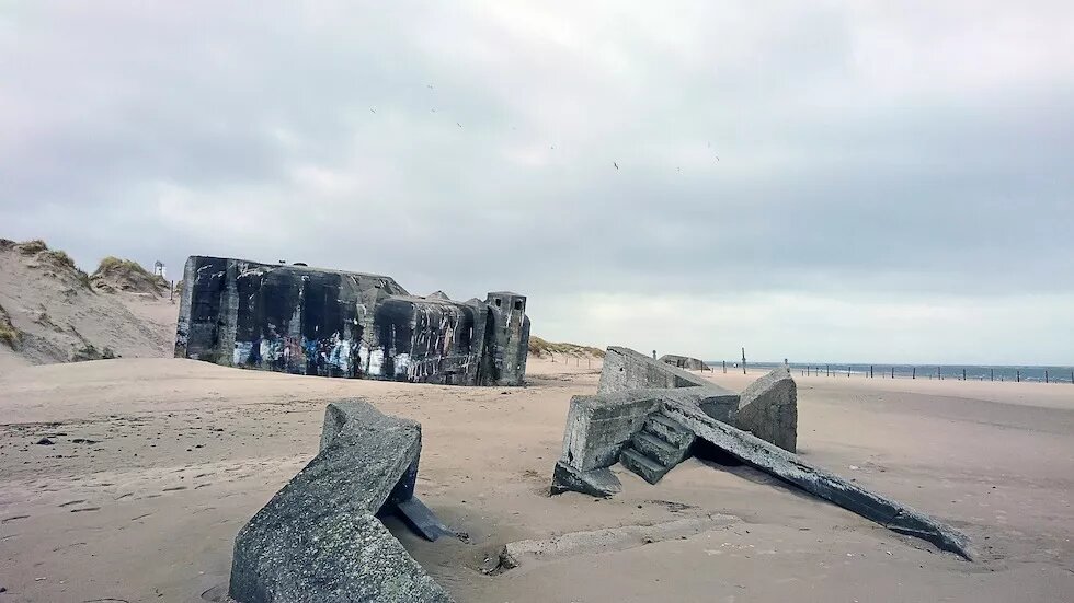 An einem Strand liegen zum Teil im Sand vergrabende Überreste eines Betonbunkers.