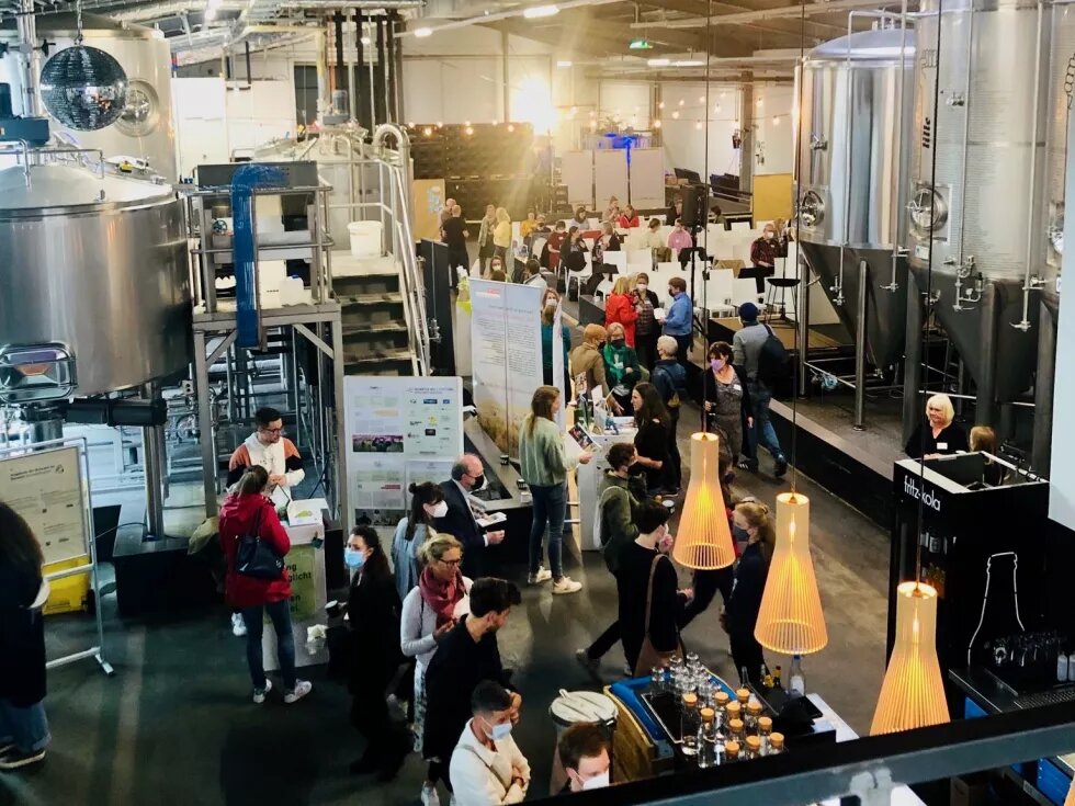 Man sihet eine große Halle, in der Braukessel für Bier stehen. Zwischen den KEsseln stehen Stände und Plakate, an denen viele Menschen stehen. Außerdem gibt es ein Buffet.