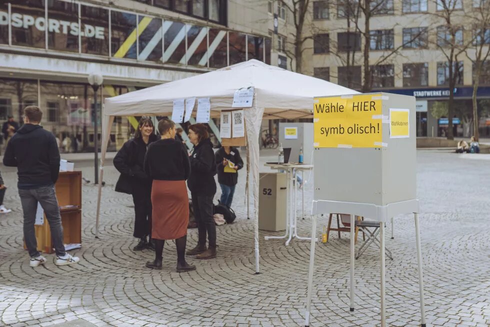 Auf einem Platz steht vorne eine Wahlkabine, auf der steht: Wähle hier symbolisch. Dahinter ist ein Pavillon aufgebaut, vor dem eine Gruppe von Menschen steht.