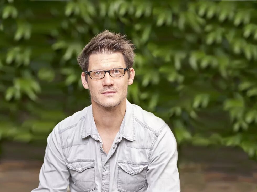 Porträt von Torsten Schäfer. Ein mittelalter Mann sitzt vor einem grünen Strauch. Er trägt ein graues Jeanshemd und eine Brille. Er hat kurze braune Haare.