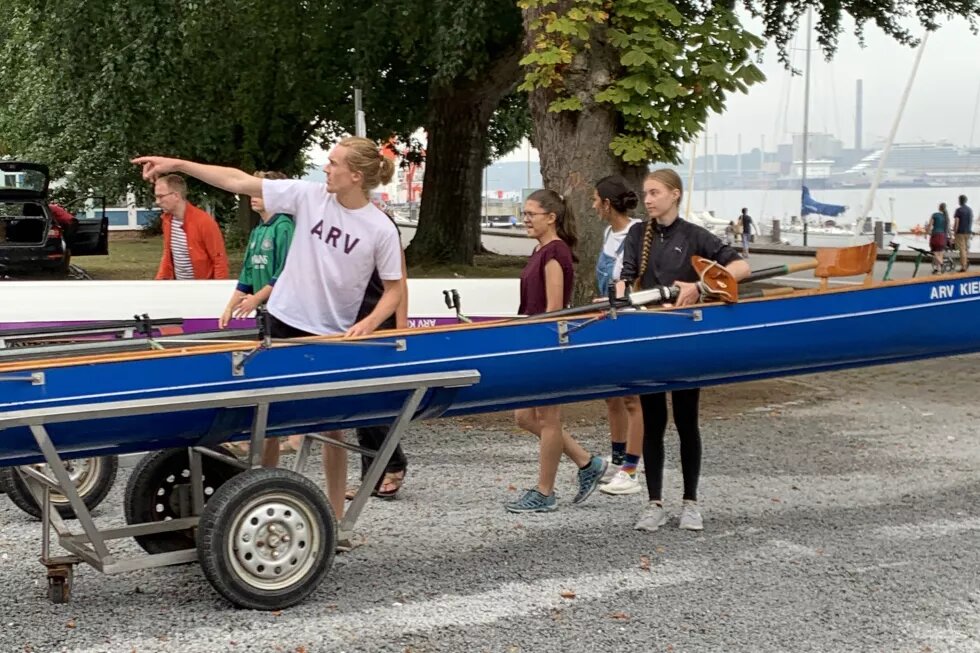 Mehrere Personen stehen um ein Ruderboot herum. Eine Person deutet nach links. Im Hintergrund sieht man Wasser und Hafen.