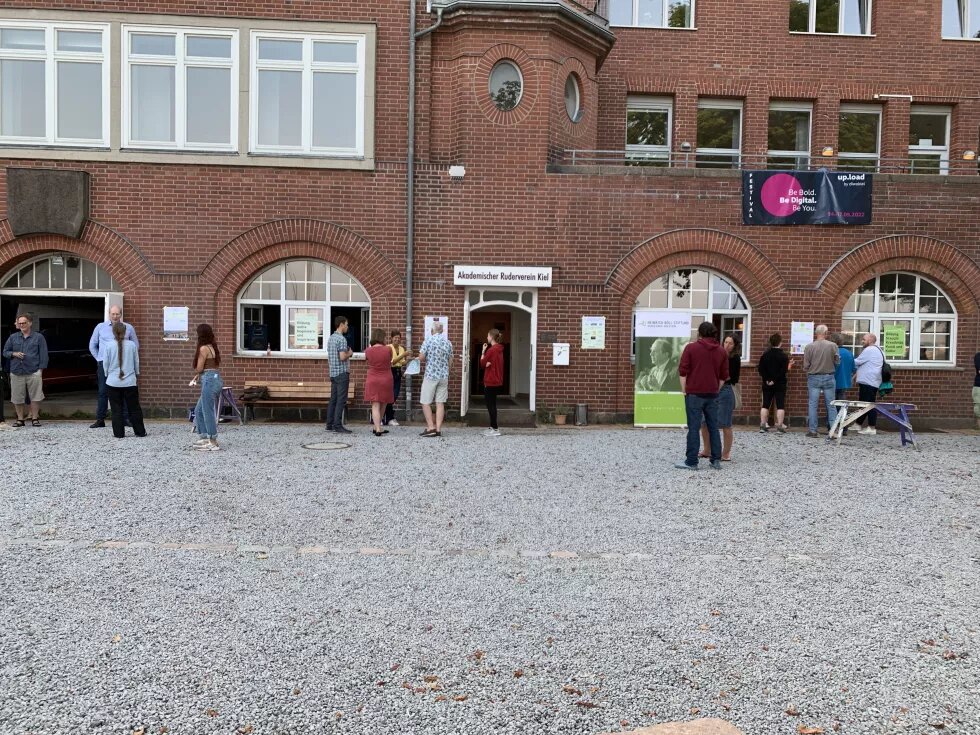 Mehrere Leute stehen vor einem Backsteingebäude. Neben dem Eingang steht ein Plakat der Heinrich-Böll-Stiftung Schleswig-Holstein.