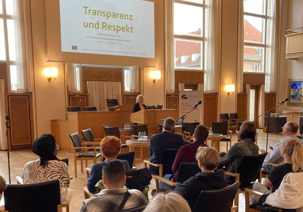 Man sieht sitzendes Publikum von hinten. Vorne steht am Podium eine blonde Person. Dahinter ist Transparenz und Respekt an die Wand projiziert.