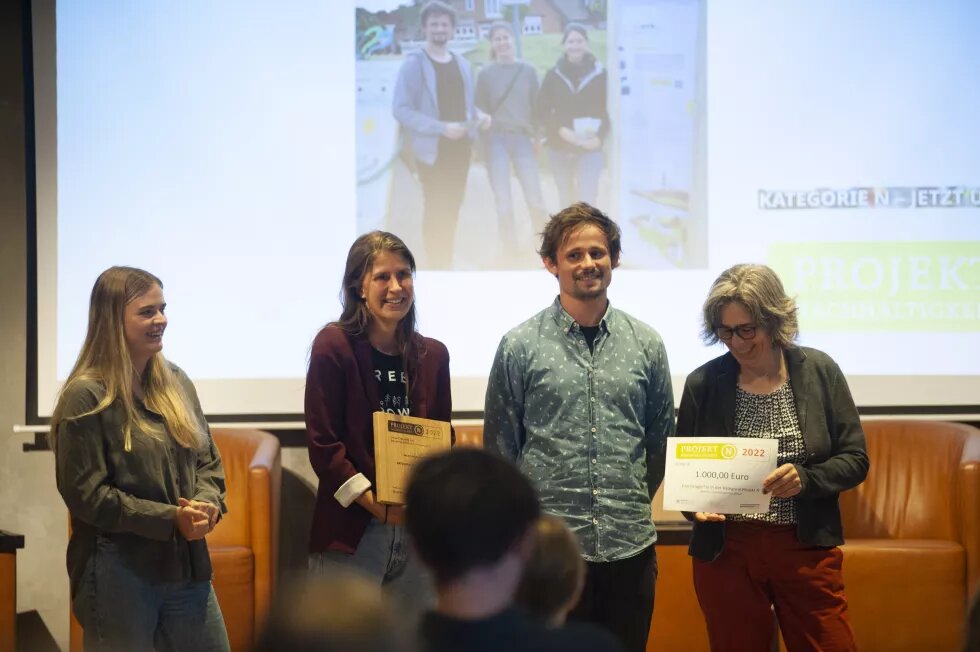 Vorne stehen viere erwachnsene Personen, die eine Urkunde in der Hand halten. Im Hintergrund ist ein Foto an eine Wand projiziert.