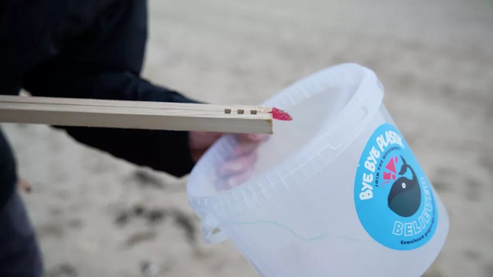 Auf einem Eimer steht "Bye bye Plastik" Mit eine Holzzange tut jemand Müll vom Strand in den Eimer.