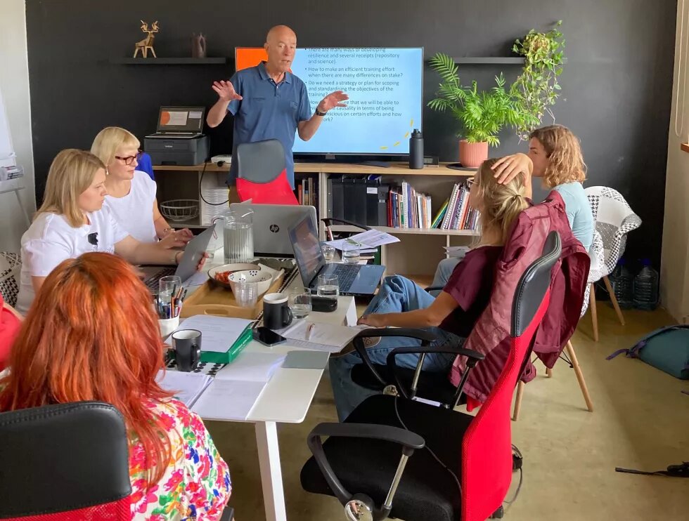 Fünf Teilnehmende vom Partnertreffen BSR Cultural Pearls in Tallinn sitzen um einen Tisch, auf dem Arbeitsmaterialien liegen. Hinten ist ein Bildschirm mit einer Präsentation, vor dem eine Person steht. 