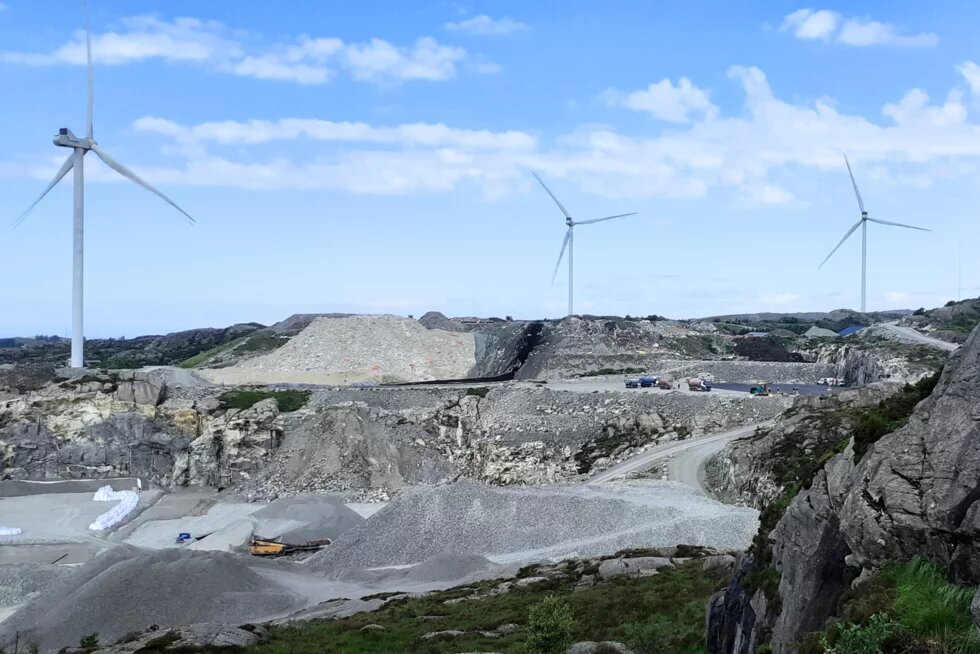 Das Bild zeigt mehrere Windkraftanlagen auf Hügeln in einer felsigen Landschaft. Im Vordergrund befinden sich große Mengen an Schotter und Kies, sowie einige Lastwagen und Maschinen. Der Himmel ist blau mit wenigen weißen Wolken.